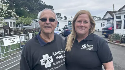 A man and woman wearing branded T-shirts stand in front of gates at the entrance to an animal sanctuary.