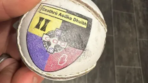 A close-up of a man's hand holding a sliotar or hurling ball embellished with the black, yellow, purple and red crest of Carryduff GAC