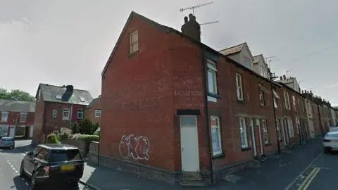 Google Streetview Google Streetview image of a road of red-brick terraced houses near Ecclesall Road in Sheffield, with parked cars outside the house and double yellow lines to the right. The front door is up two steps. There is white graffiti and a window on the wall on one side of the house, and two windows on the other wall with the Neill Road street sign.A chimney and aerial is on the roof.