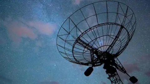 A radar disc pointed a dark night sky with an array of stars and clouds, tinged in purple, visible. The radar itself is in silhouette