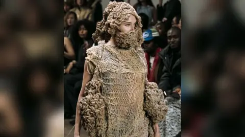 Gabrielle Thomas A model walking down a catwalk in front of a large crowd. She is wearing a dress made from brown knitted rags which covered her face, and large clumps of wool on her hips.