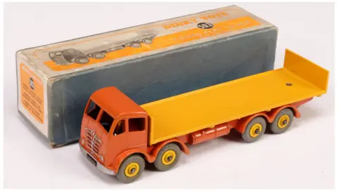 Vectis Auctions A long, orange and yellow toy truck is placed beside its cardboard box. The cab is burnt orange and has holes for windows and grey detailing depicting the grill and headlights. The wagon in the back is a single piece of yellow metal, with an uplifted section at the end. The box is yellow and grey. Text on it reads: "Dinky Toys 503 Foden Flat Truck."