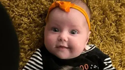 Dahlia-Rose Gartshore, a three-month-old baby girl looking at the camera, with a slight smile. She has an orange band on her head with a bow.