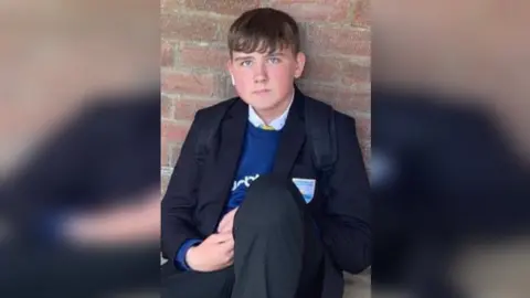 Suffolk Constabulary Harley Barfield sitting against a wall. He has short brown hair and is wearing a school uniform, including a blue jumper, with a black pack back still on his back. 