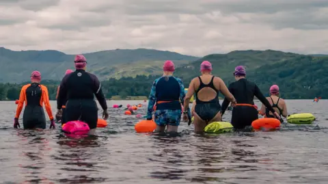 Great North Swim Seven women are walking into Windermere under a grey sky. Most are wearing wetsuits although two are just in swim suits and they are all carrying fluorescent pink, orange or yellow swim tows. The hills of the Lake District rise on the other side of Windermere. The water looks dark and the clouds beyond are forbidding.