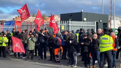 PA Media A group of around 20 people are standing in front of a bin depot. They have got red unite flags. There are also 3 police officers in the photo who are facing the protesters. You can also see onay kasab from unite the union in the crowd. Some fo the people are wearing high vis trousers but most are wearing black and darker colours with hats. 