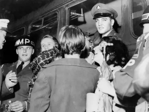 Getty Images Elvis in military uniform looking out from a train carriage. He is surrounded by women. 