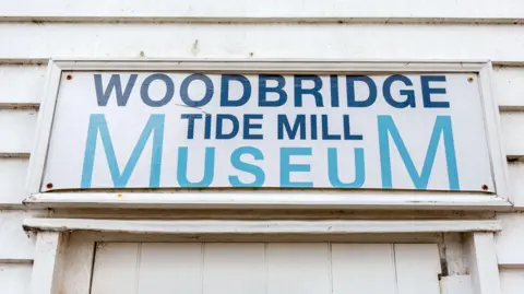 Simon Ballard A white sign is on top of a door. In blue writing it says on it 'Woodbridge Tide Mill Museum'. It is a white wooden clad building. 