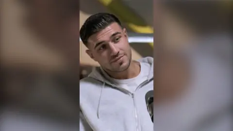 Tommy Fury has slicked back dark hair. He is wearing a white T-shirt and a grey hoodie.