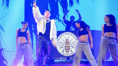 Getty Images Aitch, wearing a white fake fur jacket and black leather trousers, sings on stage at the MOBOs surrounded by four dancers wearing grey trousers and black crop tops. The stage is lined with blue strip lights and an image of the Manchester Bee appears on a DJ stand int he background