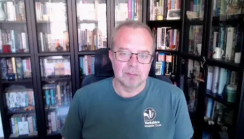 A man with short brown hair, wearing glasses and a green T-shirt, sits in a room in front of rows of bookcases filled with books of various colours.