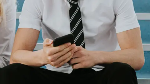 A male secondary school pupil sitting on a flight of stairs. He is holding a mobile phone and is showing it to a girl sitting next to him. He is wearing a short-sleeved white shirt and a black striped tie. Their faces are not visible.