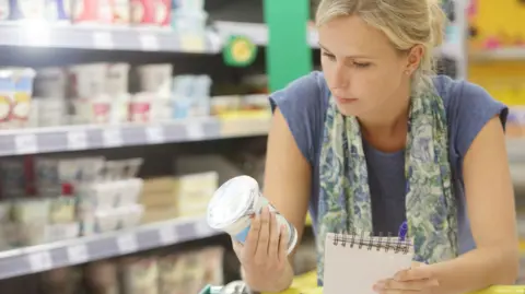 Woman with blonde hair leaning over her shopping trolley with a yoghurt pot in one hand and a notebook in the other. There is a supermarket fridge to the left of her. She is wearing a purple t-shirt and a patterned scarf.