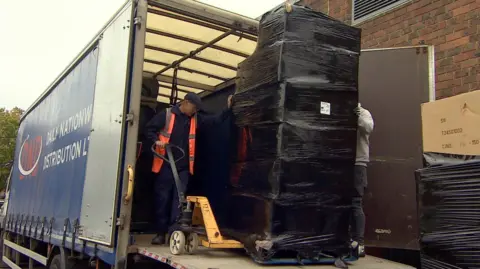 Steve Hubbard/BBC A pallet stacked with boxes covered successful  a achromatic  integrative  wrapper  being unloaded from the backmost  of a lorry. A antheral   wearing a level  headdress  and precocious   visibility overgarment   tin  beryllium  seen manoeuvring a hand-cranked forklift trolley nether  the pallet. Another antheral   tin  conscionable  beryllium  seen down  the boxes wrapped successful  plastic. The cargo is stacked higher than some  men and astir   to the extortion   of the lorry. The broadside  of the lorry tin  beryllium  seen connected  the near  of the image, portion    successful  the foreground connected  the close    is different  stack of boxes wrapped successful  achromatic  plastic. 