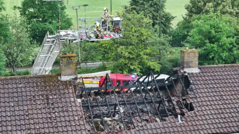 Eddie Mitchell A fire crew on an aerial ladder looking over a damaged section of the roof of a house. The timbers in the roof are badly charred, and there is rubble in the hole caused by the damage.