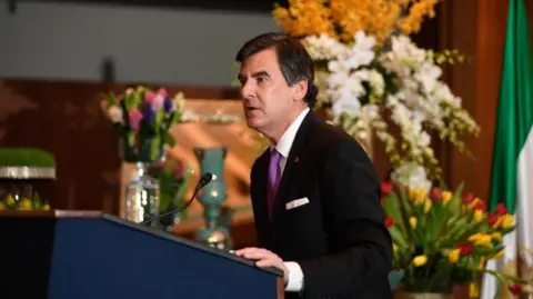 Getty Images Man wearing a suit and tie standing at a podium with his hand on the podium speaking into a microphone. In the background several bunches of flowers.