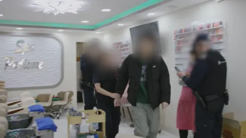 Police officers walk andocumented worker through a nail bar in Belfast after arresting him
