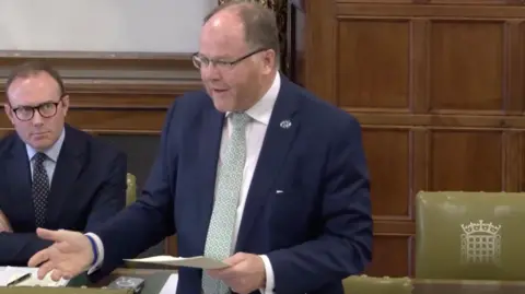 Parliament MP George Freeman addresses MPs in the House of Commons. He is wearing a blue suit and green tie.