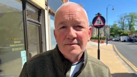 A head and shoulders shot of Robert Clarke, wearing a green pullover jumper and a light blue T-shirt. He has very short white hair and is clean shaven, standing outside on the street on a sunny day.