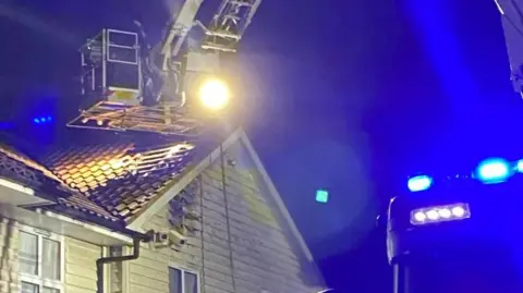 An Aerial Ladder Platform perches just above the roof of a hotel building, by which point some of its slats have been taken off so firefighters can check for hotspots. There are bright blue lights to the right of the image which are coming from the top of a fire engine. 