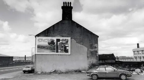 Ian Beesley A black and white photo of a car parked in front of the gable end of a house with a beer advert on its side.