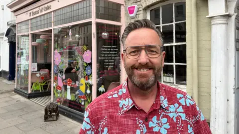 Chris Penk is standing outside House of Cake in Wellington. He is wearing a bright floral shirt and glasses.