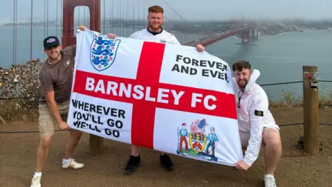 Ben Wade Three men stood with an England flag featuring a Barnsley crest with the Golden Gate Bridge in San Francisco behind them.
