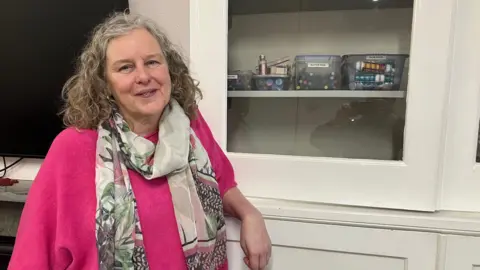 Amy Holmes/BBC A picture of Claire Holcombe from Women's Aid in Luton. She is wearing a pink jumper and has a scarf with a range of colours on it and some plants. She is stood next to a cupboard which has pens in it.