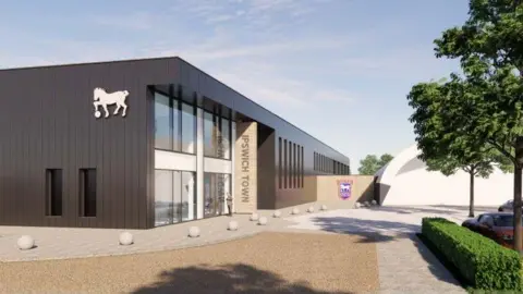 Ipswich Town Football Club Modern brown two-storey building with glass entrance frontage. There is a logo on the side of the building featuring a Suffolk punch horse with a football. There are small stone circles on the ground. The existing indoor dome is visible to the right, behind a tree and hedge.