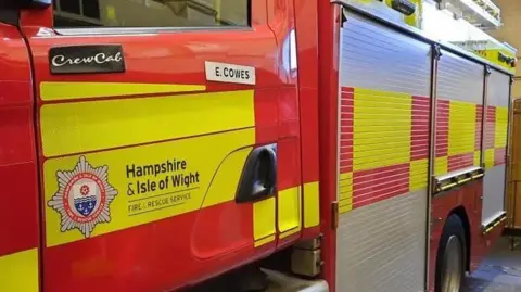 The side of a red and yellow Hampshire and Isle of Wight Fire engine. A plaque on the side says E. Cowes.