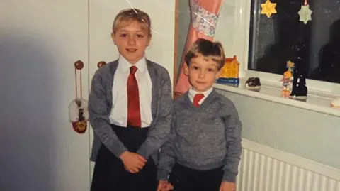 The childhood photo of Claire and Peter which was sent to Dalibor Sentic in the care package he recieved in 1992. Claire and Peter are photographed in school uniforms, stood in what appears to be a bedroom. Claire is taller than Peter and stood to the left of the image. She is wearing a black skirt, white blouse, red tie, and grey cardigan. She has blonde hair with a fringe. Her hair is tied up. Peter is shorter than Claire and is photographed to the right of her. He is wearing black trousers, a white shirt and red tie, with a grey jumper over the top.