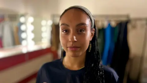MARTIN GILES/BBC A head and shoulders shot of Miah Robinson. She has a slight smile and is looking at the camera. She is standing in a theatre dressing room. The background behind her is blurry, but a mirror surrounded by white lights can be seen, as well as a rack of clothes. 