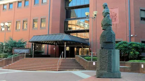 The outside of a red brick building with a glass covered entrance and a statue of several heads on top of each other outside