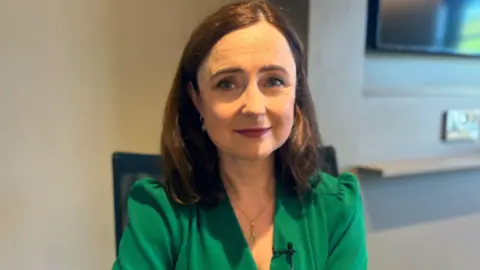 A woman with shoulder length brown hair is facing a camera. Behind her is the bottom corner of a TV and the wall is a dark cream. 