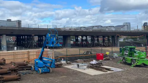 Gateshead flyover is fenced off and propped up by dozens of steel props. In front of the fencing is a green machine and a blue fork-lift type machine. They are all on dirt ground and lots of industrial parts lie on the ground.