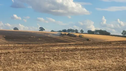 Firefighters at the scene, with what appears to be a burnt out combine harvester visible in the shot.
