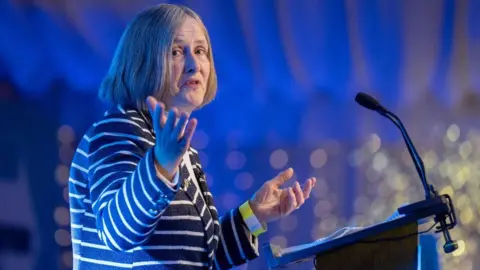 Helen McDade, who has short grey hair, speaks at a podium while illuminated in blue lighting 