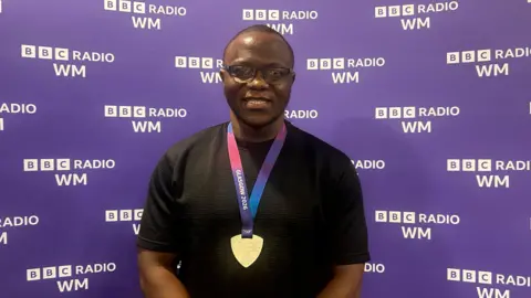 A man wearing glasses, a black short-sleeved textured shirt and a silver Glasgow 2026 medal on a purple and blue ribbon poses in front of a purple BBC Radio WM branded backdrop.