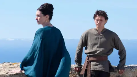 Tom Holland and Anne Hathaway in a film still from the film The Odyssey. Tom Holland is leaning against a wall with a scenic greek sea view behind him. He is wearing a dark olive green shirt and dark trousers. Anne hathaway is looking to the left and wearing a flowing flowing blue dress