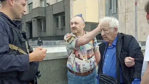 Handout An elderly man, with grey hair, a suit jacket and blue shirt, is frowning, while two men put their arms through his, holding him. The man on the left of him has a boldly-patterned shirt and moustache, and is talking while gestring to the elderly man. A police officer in navy uniform is looking at both of them.