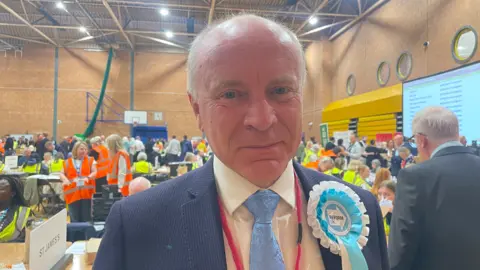 A man is wearing a blue jacket and tie, a light shirt and light blue and white rosette. He is close to the camera and many people are inside behind him in the background.