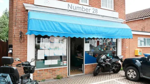 John Fairhall/BBC The outside of Number 28 community centre. There is a mobility scooter, a motorbike, and a car parked outside. It has a blue canopy above its single-door at the front.