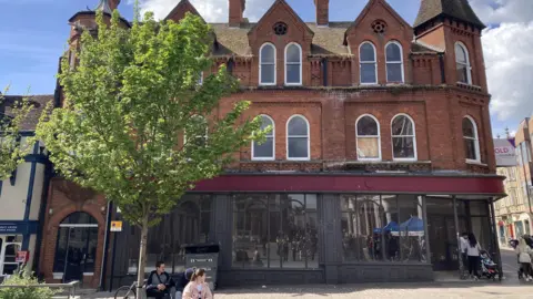 The Grimwades stores in Ipswich. It is a three-storey red brick building. The lower storey shows an empty shore with large windows and a large entrance. The two upper floors have numerous windows. A large tree sits outside the building. People can be seen walking nearby and sat on benches nearby.