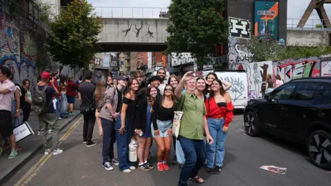 PA Media A group of people take a selfie on Brick Lane with the Banksy work in the background