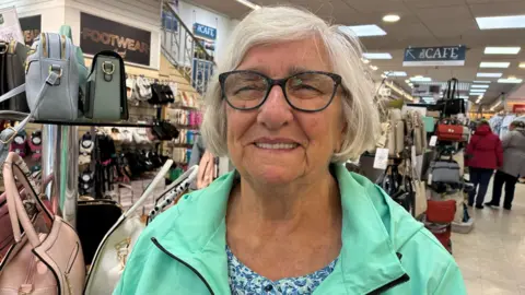 Patricia is grey haired and is smiling. She has glasses and is wearing a teal raincoat. She is stood inside the shop.