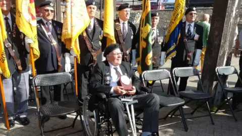 BBC Roy Mills in a wheelchair dressed in a suit and an army cap with his medals on the lapel. Behind him six men are in similar dress, holding large yellow regimental flags.