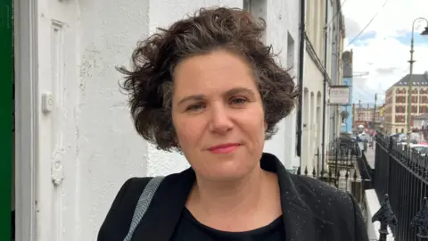 BBC Claire Hanna on a busy street wearing a dark suit. Behind her is a white wall, and a green door. To her right are rows of black railings 