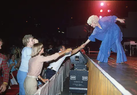 Ira Berger / Alamy At a live concert, country music superstar Dolly Parton goes to the front of the stage to shake hands with fans. She wears a purple and bedazzled flowy top and pants.