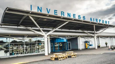 The main entrance to Inverness Airport. The name of the airport is spelled out in large lettering on a metal canopy above a blue revolving door.