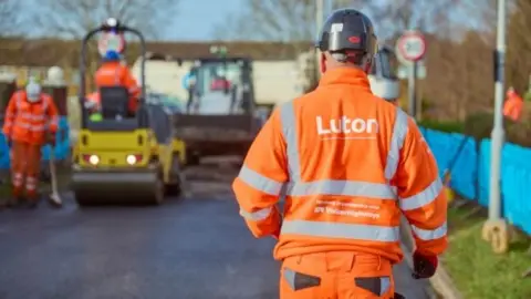 Luton Council A man wearing bright orange high-vis clothing is walking towards some roadworks ahead of him. On his back the words "Luton" are written in white font.